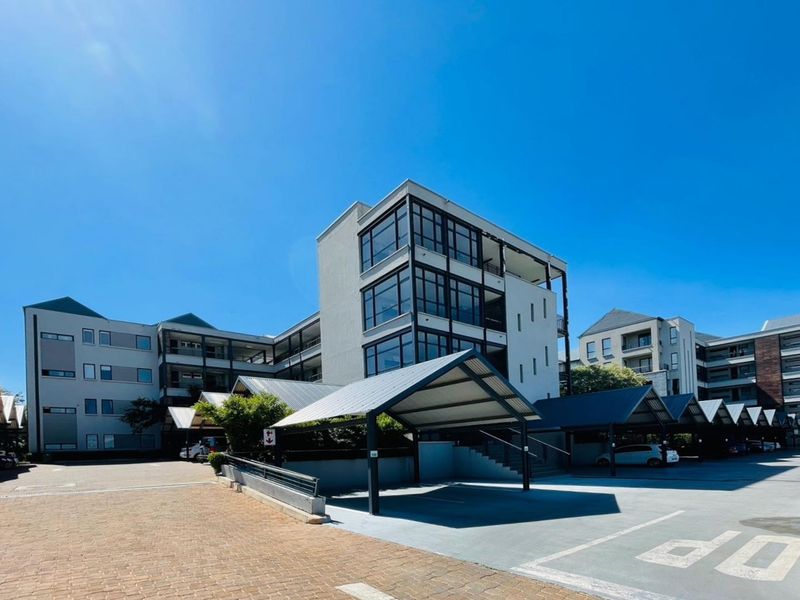 Exterior view of a modern apartment complex featuring covered parking bays, paved driveway, and multi-story residential buildings with large glass windows.