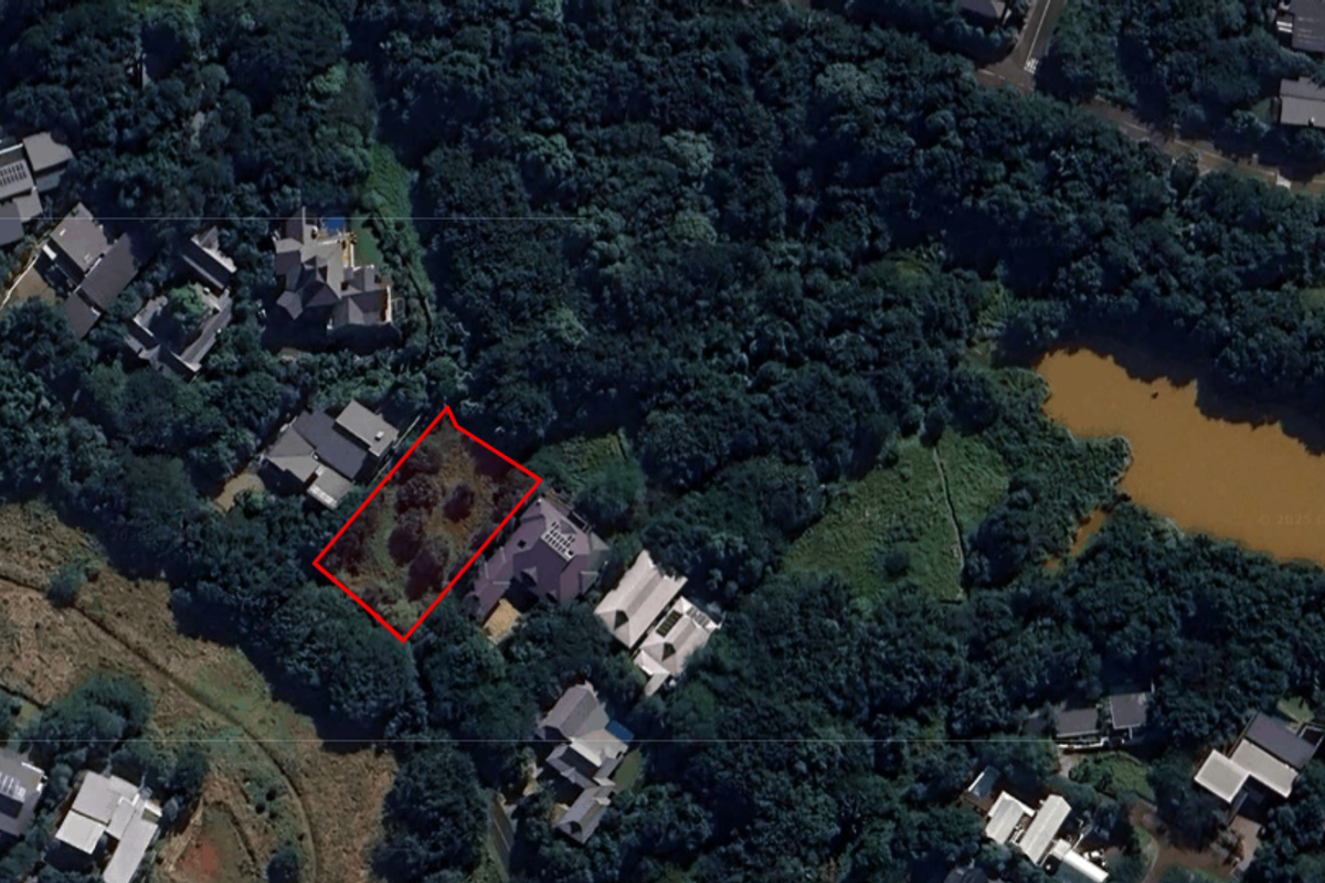An aerial view of a vacant residential plot highlighted in red, situated within a lush, wooded estate environment near a small lake.