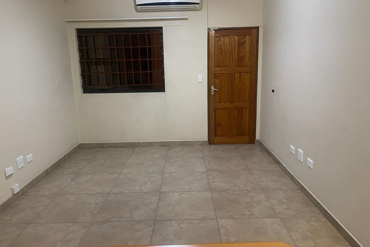 An empty bedroom featuring tiled flooring, a wall-mounted air conditioning unit, a window with security bars, and a wooden door.