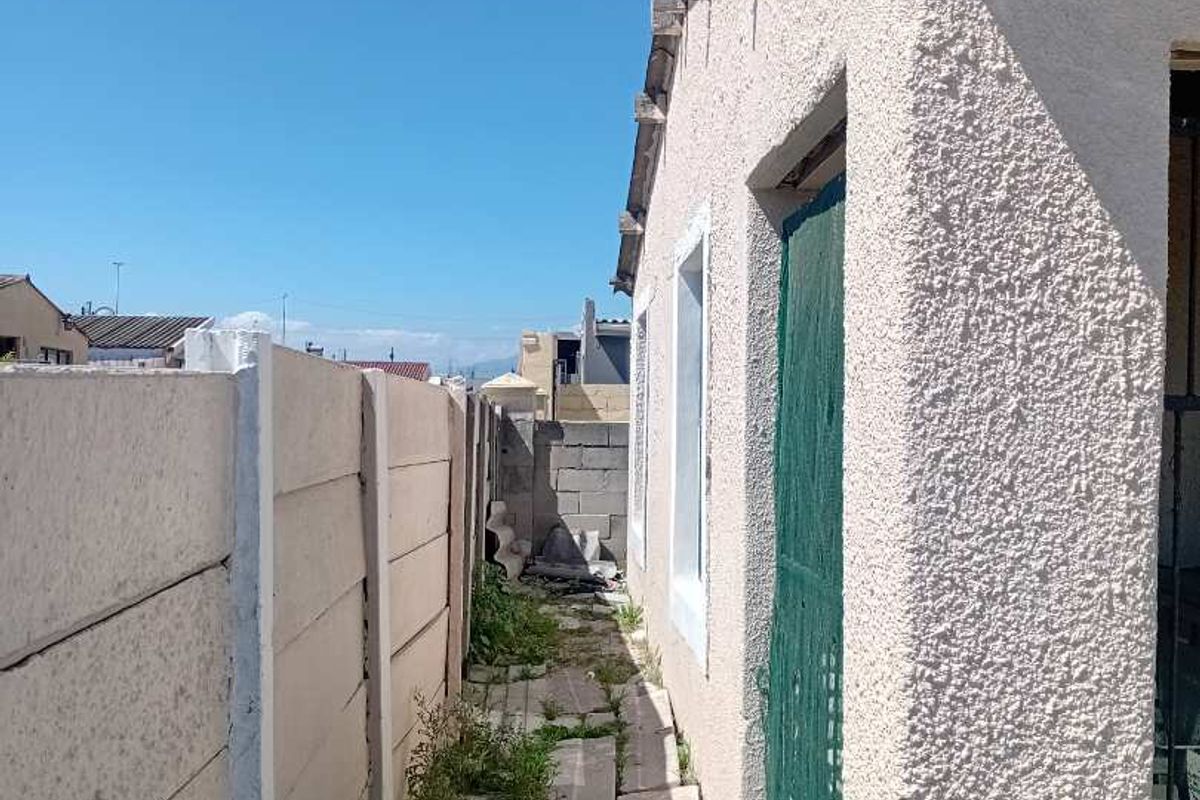 Narrow side passage featuring a precast concrete boundary wall, textured exterior wall finish, and weathered paved walkway requiring maintenance.