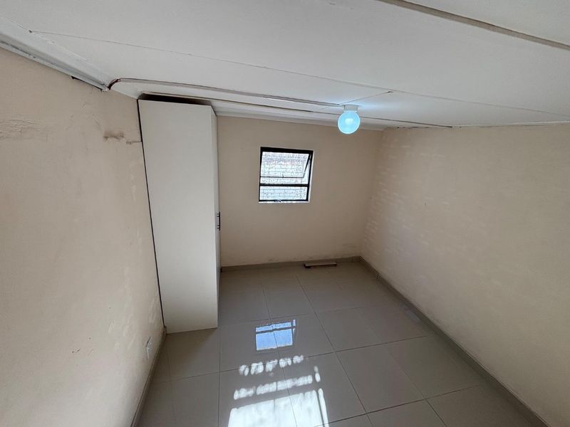 A small, empty bedroom featuring tiled flooring, a built-in wardrobe, and a single window. The room requires cosmetic updates and ceiling repairs.