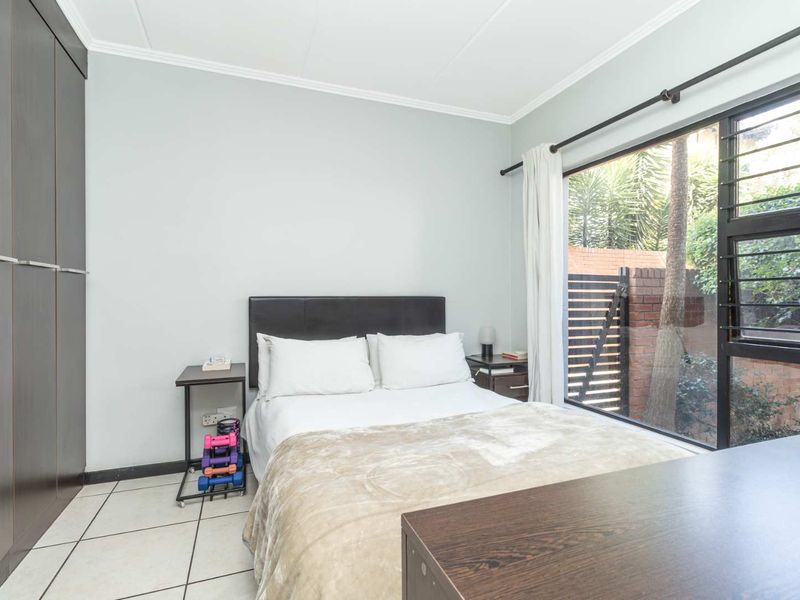 A well-lit bedroom featuring built-in cupboards, tiled flooring, and a large window overlooking the exterior garden area.