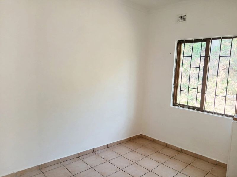 A clean, empty bedroom featuring neutral tiled flooring, a window with security bars, and a wall-mounted ventilation grille.