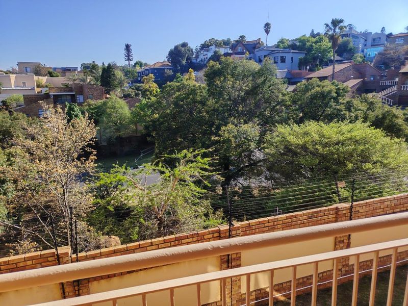 Elevated view overlooking a lush residential neighborhood with mature trees, featuring a brick boundary wall with integrated electric fencing and a metal balcony railing.
