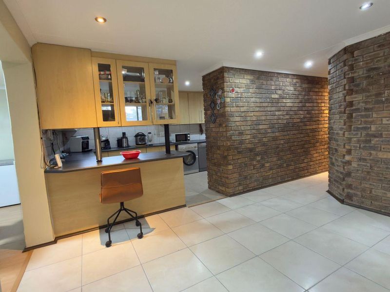 A functional kitchen featuring light wood cabinetry, laminate countertops, and a distinctive exposed brick feature wall, complemented by modern recessed lighting and tiled floors.