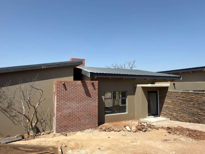 Modern single-story home exterior featuring a combination of brick, stone cladding, and corrugated metal roofing with an unfinished surrounding landscape.