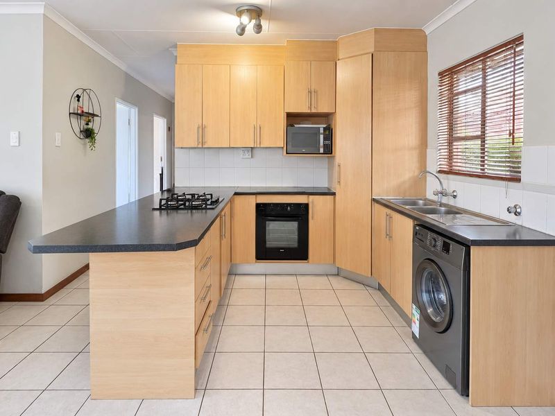 Functional kitchen featuring light wood cabinetry, dark laminate countertops, built-in gas hob, oven, and integrated laundry plumbing, finished with neutral floor tiles.