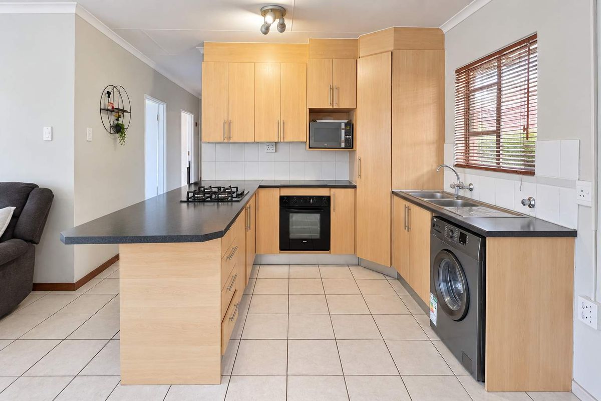 Functional kitchen featuring light wood cabinetry, dark laminate countertops, built-in gas hob, oven, and integrated laundry plumbing, finished with neutral floor tiles.