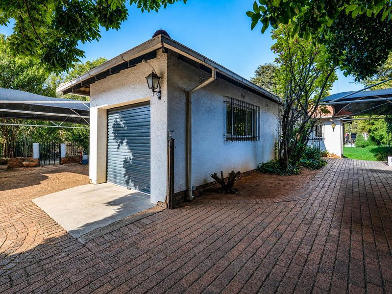 A detached single garage featuring a roller shutter door, set within a paved courtyard area with established trees and security features.
