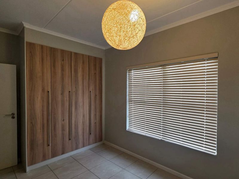 A clean, empty bedroom featuring modern wood-finish built-in cupboards, neutral tiled flooring, and venetian blinds, offering a versatile space ready for personalization.