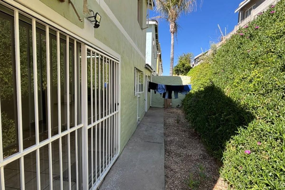 A narrow rear exterior walkway featuring a concrete path, security gate, and sliding door access, bordered by a landscaped hedge.