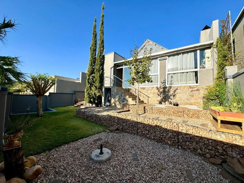 Rear exterior view featuring a landscaped garden with gabion retaining walls, stone-clad foundation, and a metal staircase leading to the elevated patio.