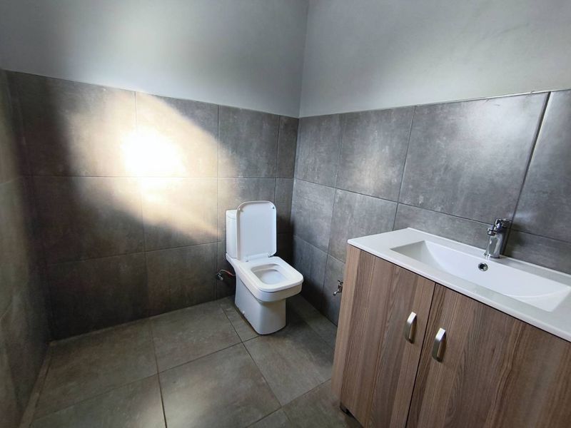 A clean, modern bathroom featuring grey wall and floor tiles, a white toilet, and a wood-finish vanity cabinet with a basin.