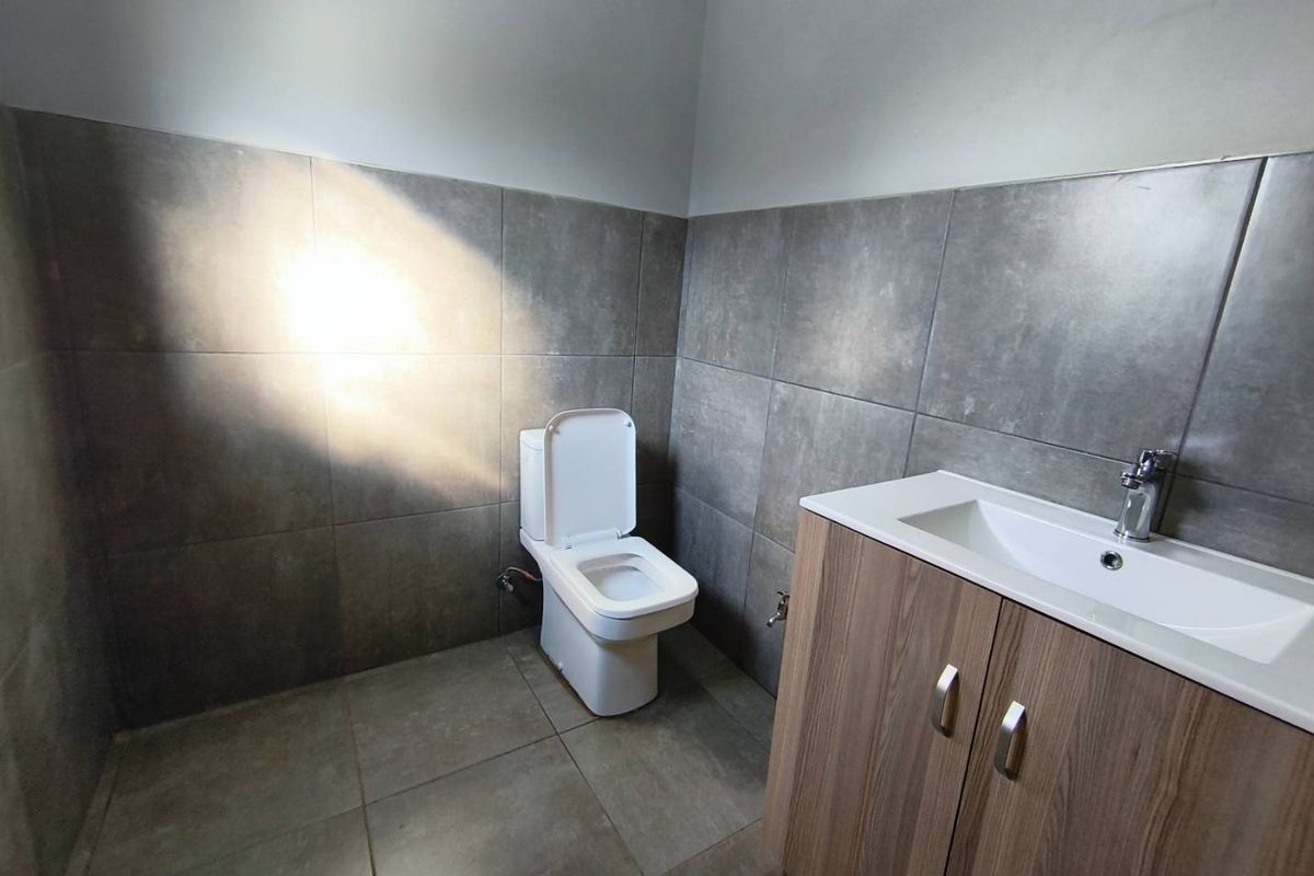 A clean, modern bathroom featuring grey wall and floor tiles, a white toilet, and a wood-finish vanity cabinet with a basin.