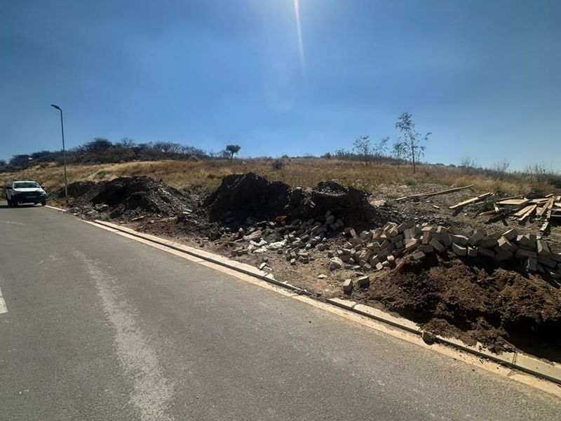 Street view of a vacant residential plot featuring an asphalt road, concrete curbing, and visible construction debris and soil piles on the site.