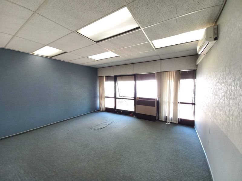A spacious, empty office suite featuring a drop ceiling with recessed lighting, wall-mounted air conditioning unit, and large windows providing natural light.
