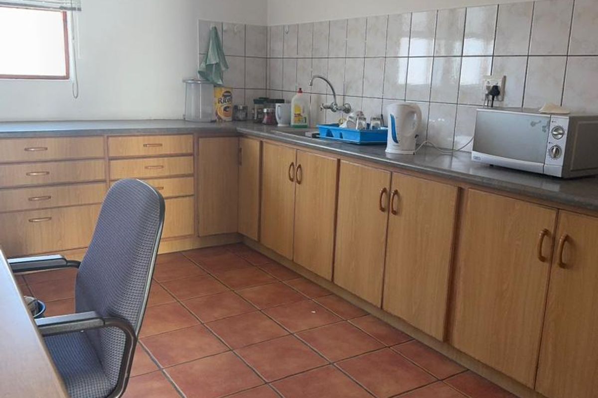 A functional office kitchenette featuring wooden cabinetry, tiled backsplash, stainless steel sink, and durable floor tiles.