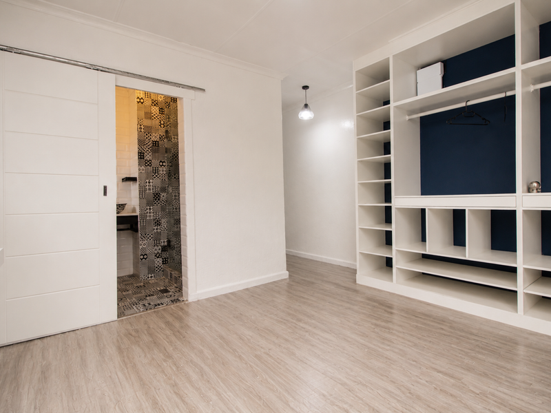 A bright bedroom featuring modern laminate flooring, a large built-in wardrobe with shelving, and a sliding door leading to an en-suite bathroom.