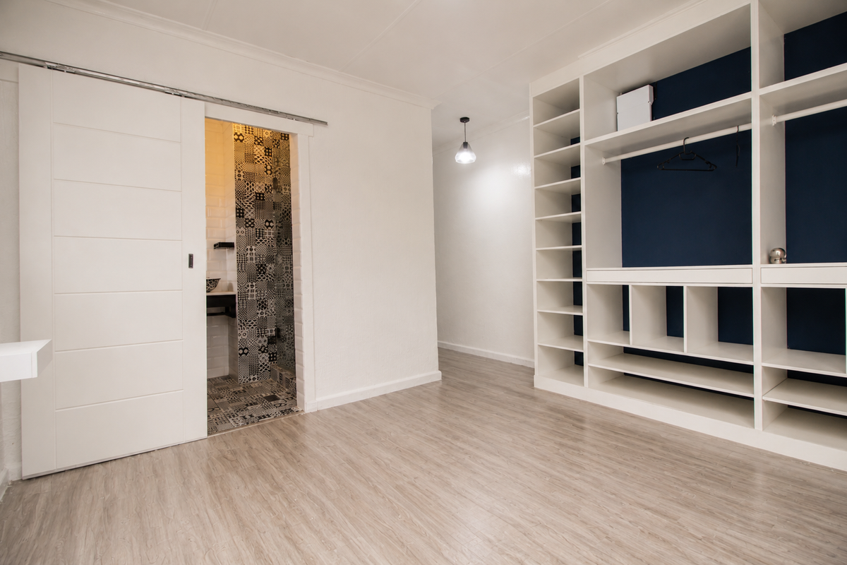 A bright bedroom featuring modern laminate flooring, a large built-in wardrobe with shelving, and a sliding door leading to an en-suite bathroom.