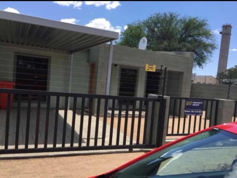 Front exterior view of the apartment featuring a paved driveway, covered carport, and secure perimeter fencing with an integrated gate.