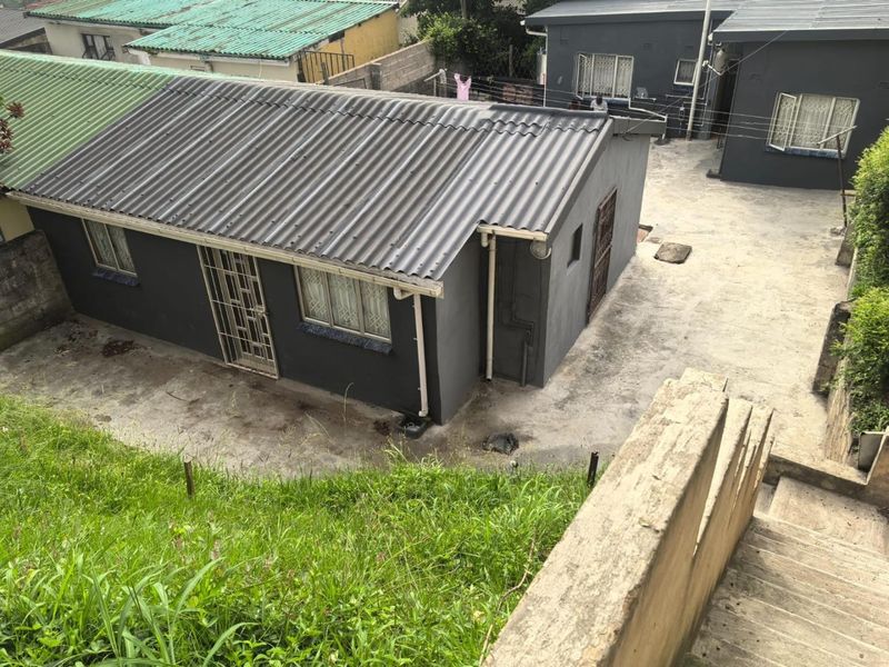 An elevated view of the rear exterior of the property, featuring a corrugated metal roof, concrete paving, and security gates on the windows and doors.