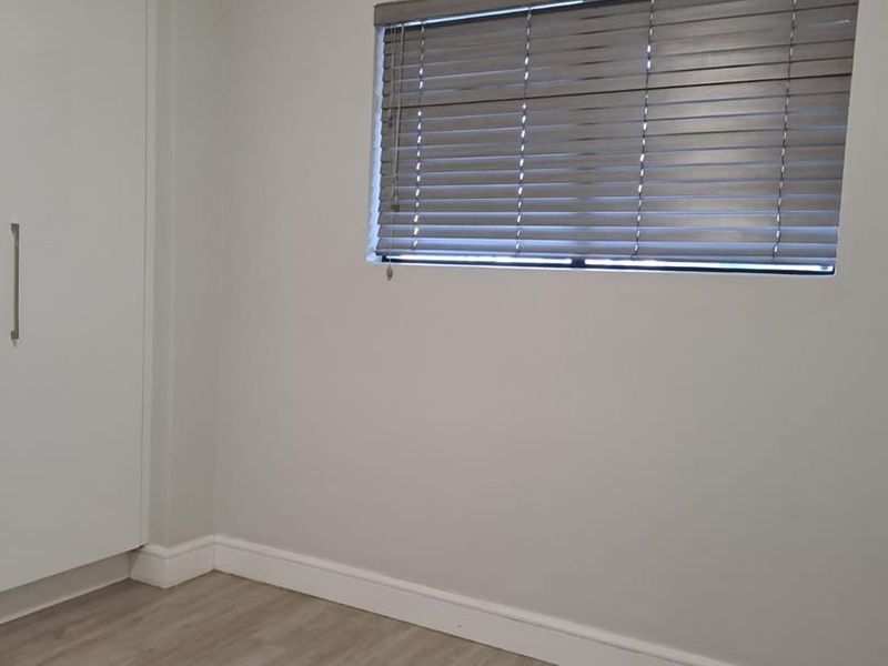 A clean, empty bedroom featuring light-toned laminate flooring, neutral walls, a built-in wardrobe, and fitted venetian blinds.