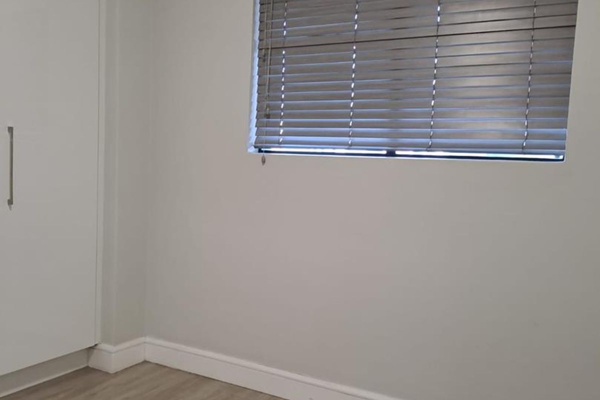 A clean, empty bedroom featuring light-toned laminate flooring, neutral walls, a built-in wardrobe, and fitted venetian blinds.