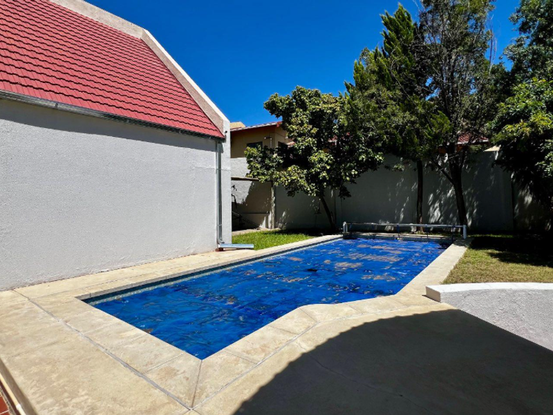 A well-maintained outdoor swimming pool area featuring a blue pool cover, adjacent to a house with a red tiled roof and boundary wall.