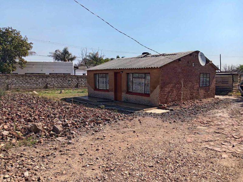 A single-story detached house featuring an exposed brick facade, corrugated roof, and security bars on windows, set on a large plot with a boundary wall.