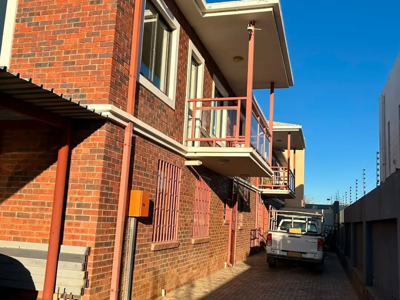 Exterior view of the apartment building featuring face brick construction, balconies with metal railings, and a paved driveway with perimeter electric fencing.
