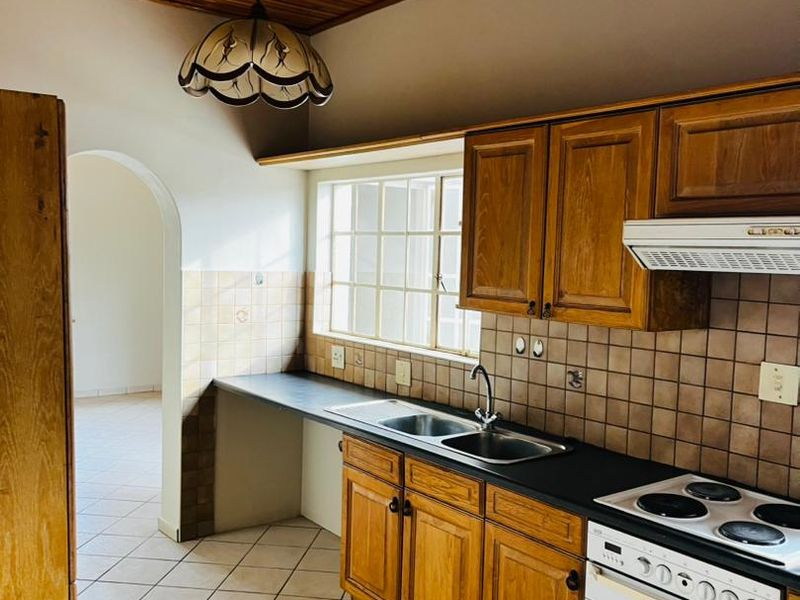 A functional kitchen featuring timber cabinetry, tiled backsplash, built-in oven with hob, and a distinctive timber-paneled ceiling, leading through an archway to adjacent living space.