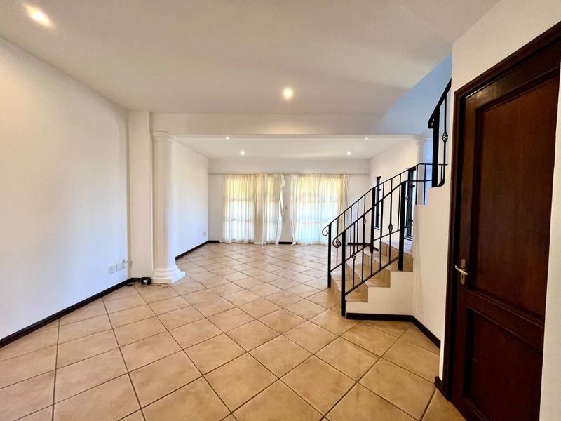 Spacious, empty living area featuring neutral tiled flooring, a decorative column, and a staircase with wrought iron balustrades, leading to a bright windowed space.