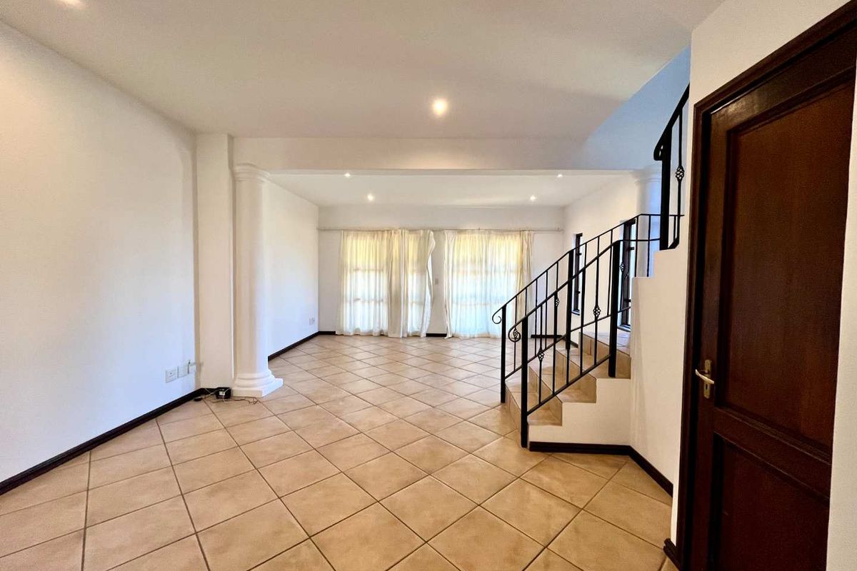 Spacious, empty living area featuring neutral tiled flooring, a decorative column, and a staircase with wrought iron balustrades, leading to a bright windowed space.