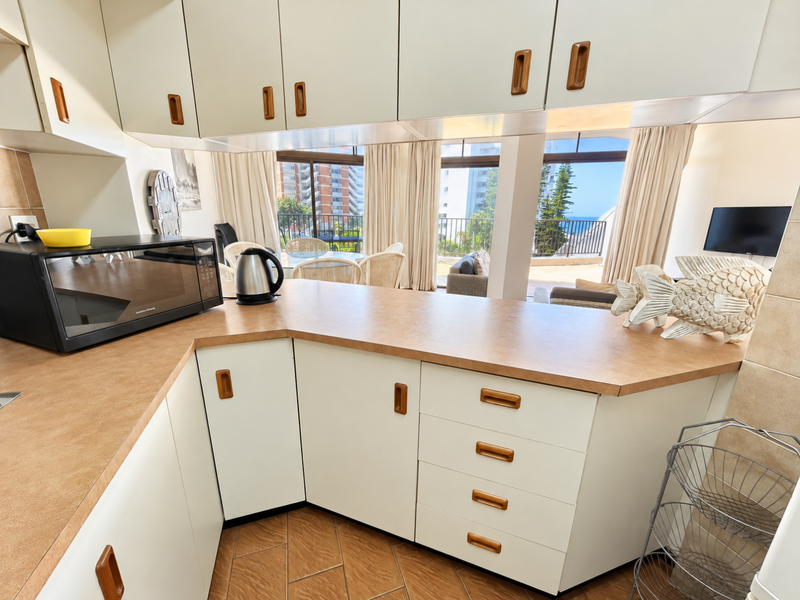 A functional kitchen featuring laminate countertops and ample built-in cabinetry, offering a view through to the adjacent living area and balcony.
