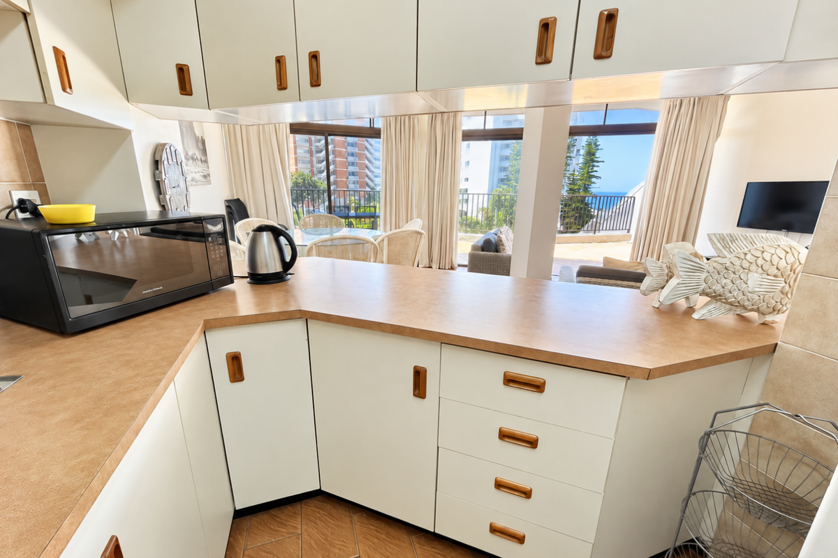 A functional kitchen featuring laminate countertops and ample built-in cabinetry, offering a view through to the adjacent living area and balcony.