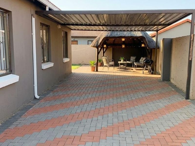 A well-maintained rear exterior area featuring a paved driveway, a covered carport, and a traditional thatched lapa entertainment space.