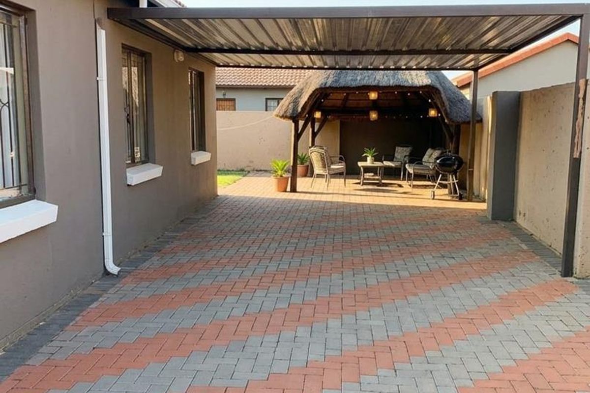 A well-maintained rear exterior area featuring a paved driveway, a covered carport, and a traditional thatched lapa entertainment space.