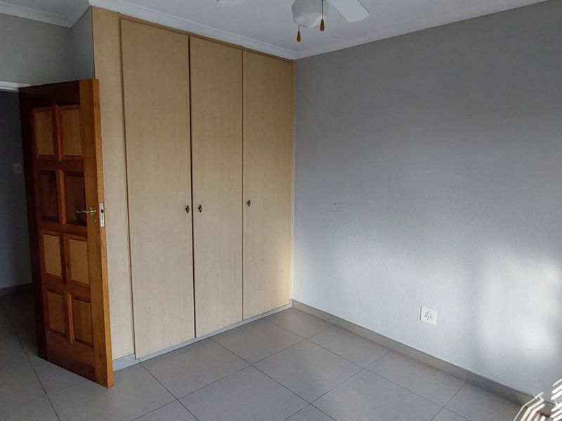 A clean, empty bedroom featuring neutral tiled flooring, built-in wooden cupboards, and a ceiling fan, with an adjacent view into a bathroom.