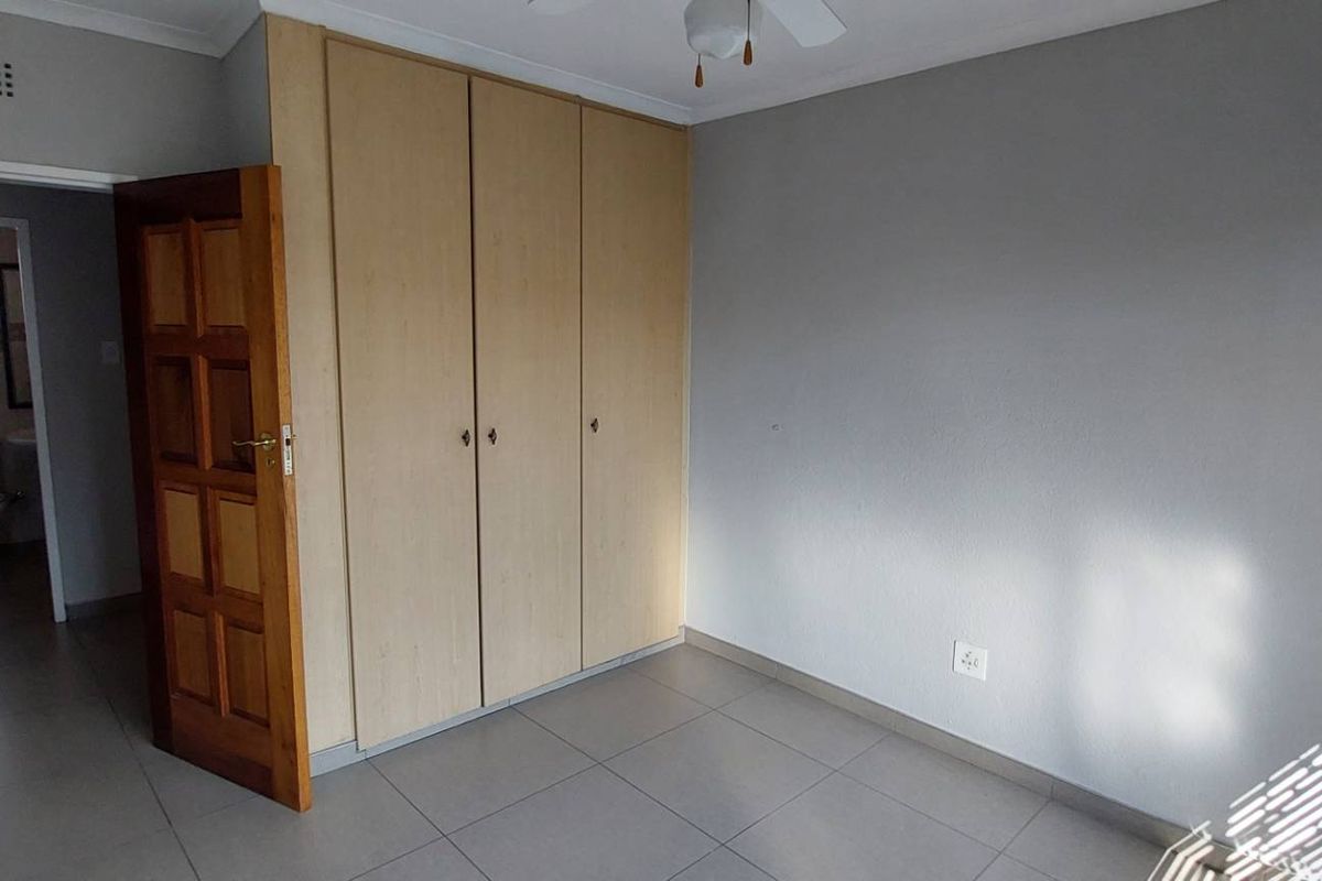A clean, empty bedroom featuring neutral tiled flooring, built-in wooden cupboards, and a ceiling fan, with an adjacent view into a bathroom.