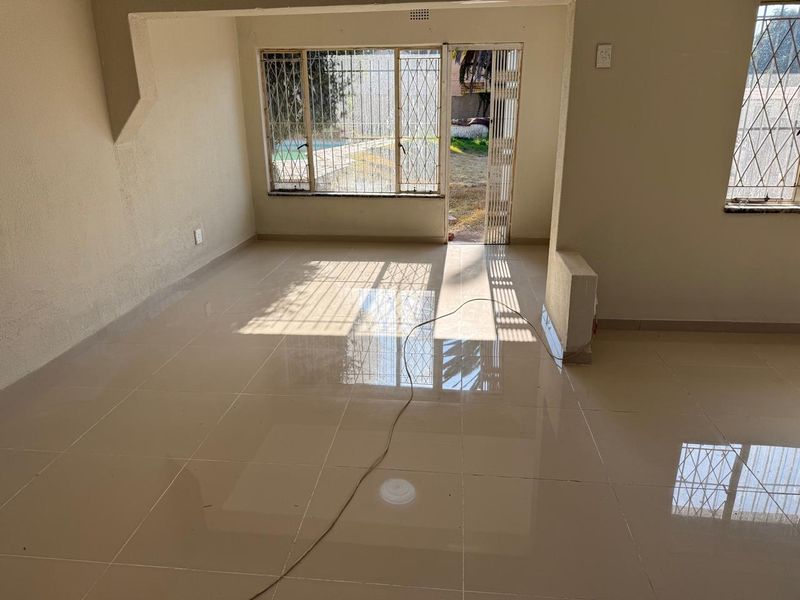A bright, empty living room featuring polished tiled flooring and direct access to the garden through a sliding door with security gates.
