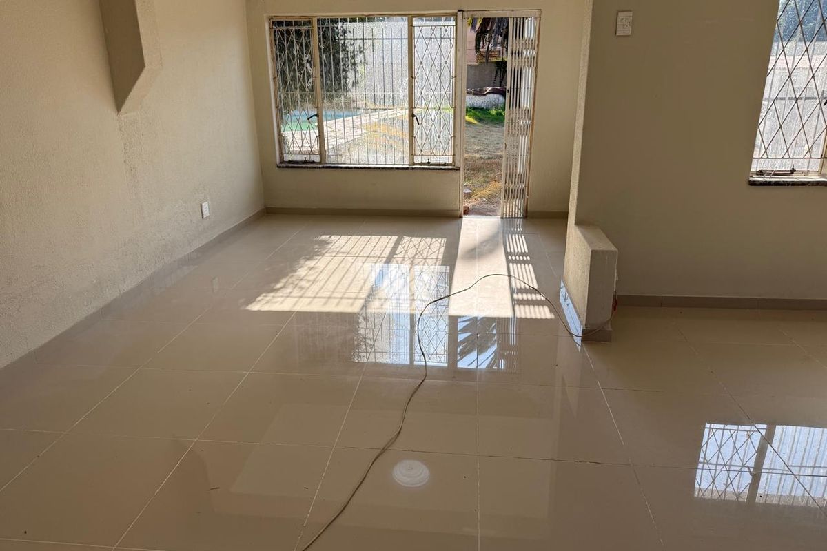 A bright, empty living room featuring polished tiled flooring and direct access to the garden through a sliding door with security gates.