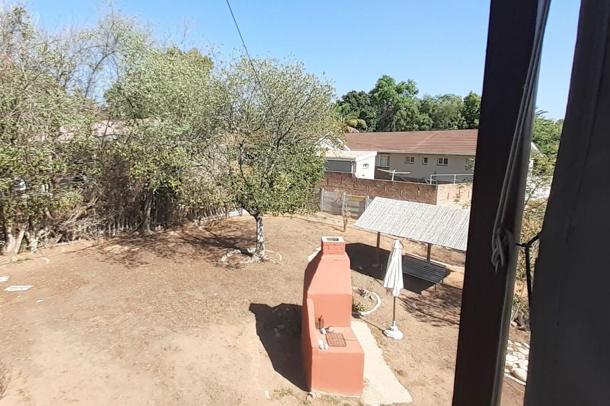 A view of the rear garden featuring a built-in brick braai, a thatched gazebo, and mature trees.