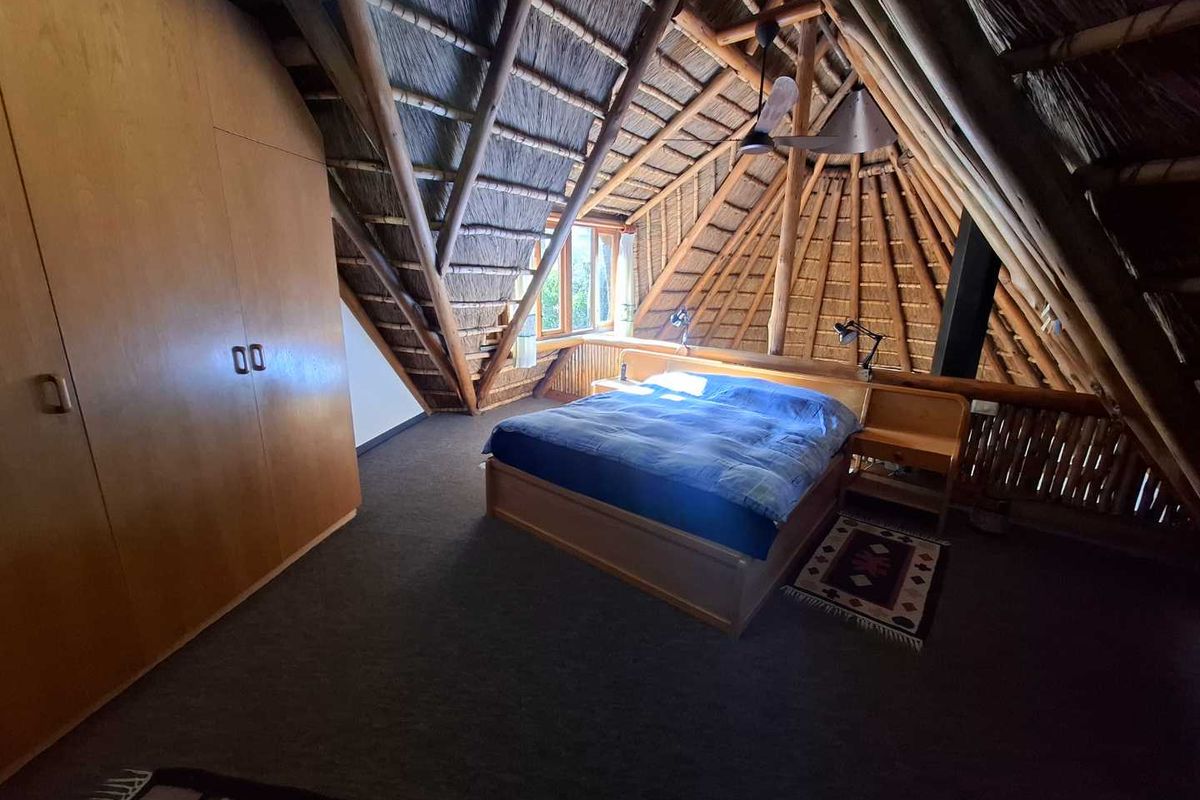 A unique loft-style bedroom featuring a high-pitched thatched roof with exposed timber beams, built-in wooden cabinetry, and carpeted flooring.