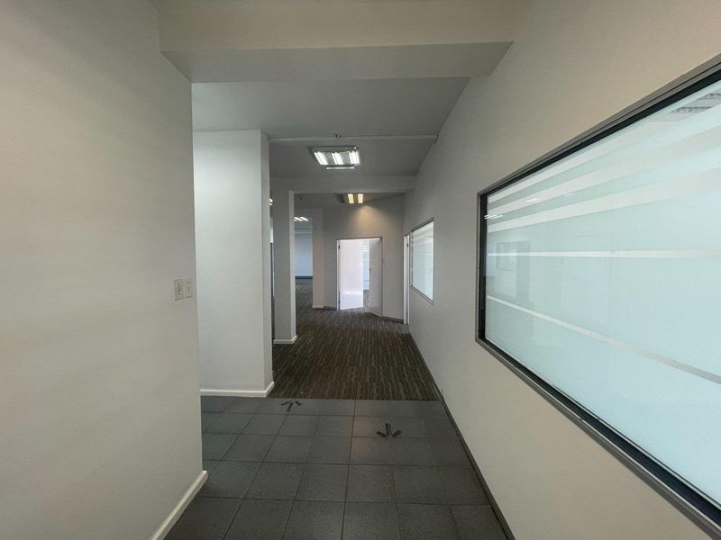 A clean, vacant office hallway featuring a mix of tiled and carpeted flooring, recessed lighting, and a frosted glass partition wall.