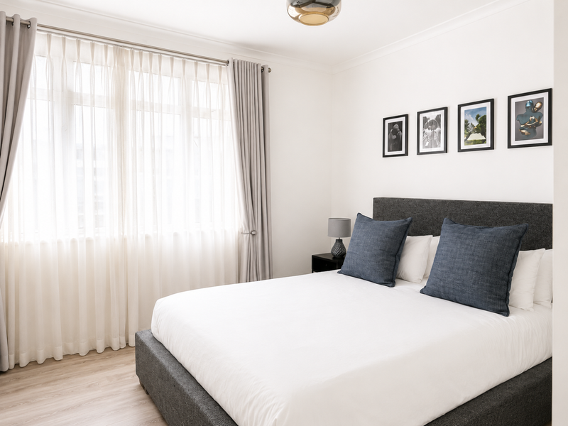 A bright and well-maintained bedroom featuring neutral walls, light wood-look flooring, and large windows dressed with elegant floor-to-ceiling curtains.
