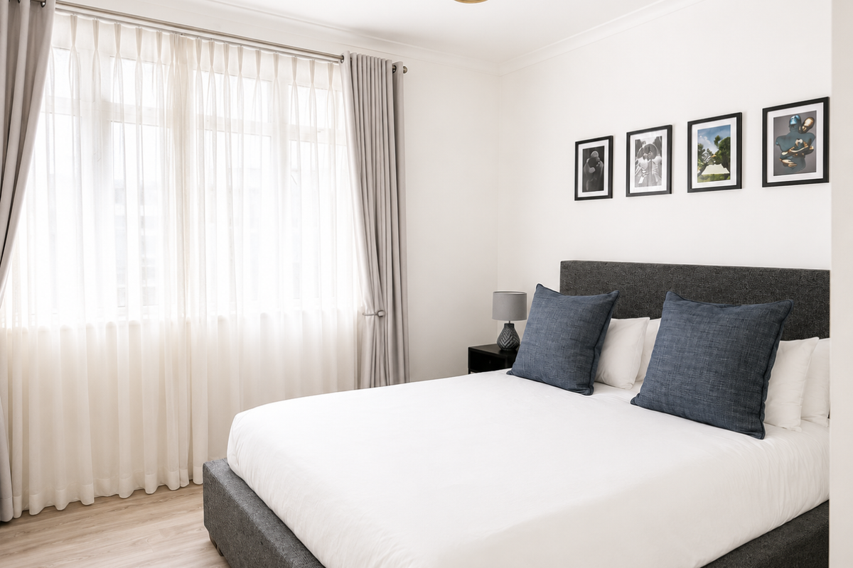 A bright and well-maintained bedroom featuring neutral walls, light wood-look flooring, and large windows dressed with elegant floor-to-ceiling curtains.