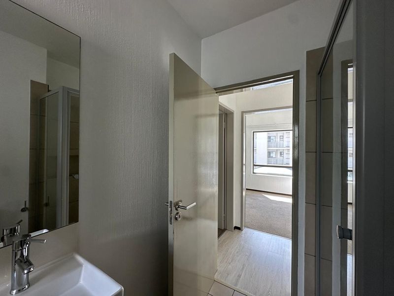 A functional bathroom featuring a vanity basin with a mixer tap, wall mirror, and a shower enclosure, leading into a hallway.