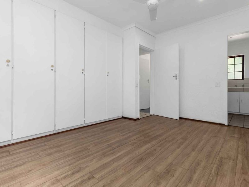 A bright, empty bedroom featuring laminate flooring, built-in cupboards, and a ceiling fan, with a view into an adjacent tiled kitchen area.