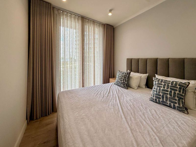 A well-appointed bedroom featuring modern recessed lighting, neutral tones, laminate flooring, and floor-to-ceiling curtains leading to a balcony.