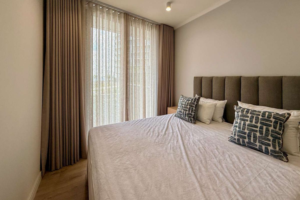 A well-appointed bedroom featuring modern recessed lighting, neutral tones, laminate flooring, and floor-to-ceiling curtains leading to a balcony.